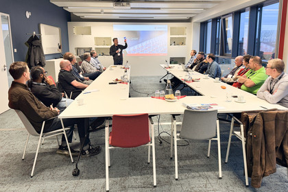 Mehrere Personen sitzen in einem Besprechungsraum an einem großen U-förmigen Tisch, während eine präsentierende Person vorne am Bildschirm steht und auf eine Folie zeigt.