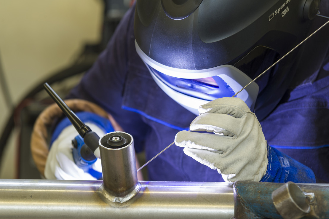 Eine schweißende Person mit Helm und Handschuhen führt präzise eine WIG-Schweißung an einem Edelstahlrohr durch, das eine T-Verbindung mit einem Stutzen aufweist.