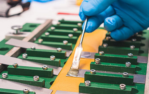 Hände in blauen Handschuhen tragen Kleber auf eine grüne Leiterplatte mit elektronischen Bauteilen.