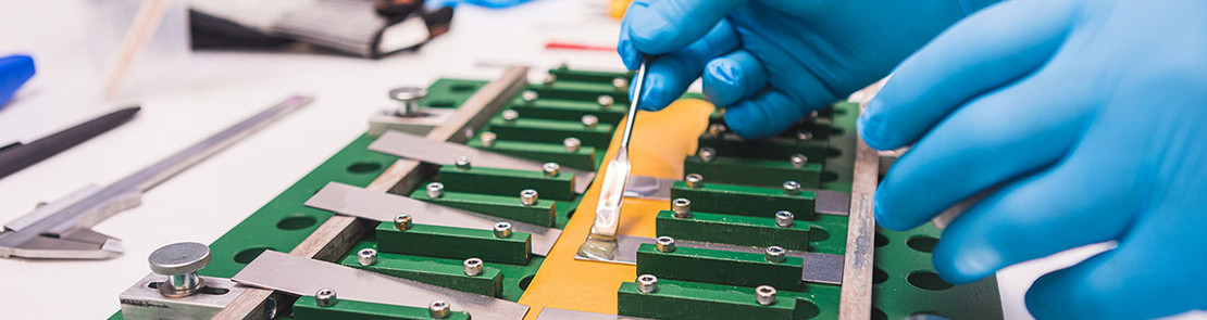 Hände in blauen Handschuhen tragen Kleber auf eine grüne Leiterplatte mit elektronischen Bauteilen.