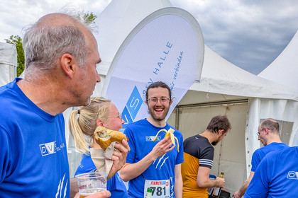 Zwei Männer und eine Frau in blauen SLV Halle T-Shirts essen und trinken vor einem Zelt mit SLV Halle Logo, sie lachen und genießen die Pause.