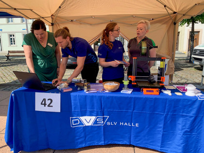 Vier Personen stehen an einem Messestand unter einem Pavillon mit Tischdecke der DVS SLV Halle, auf dem ein 3D-Drucker, Werkstücke und ein Laptop präsentiert werden; Tischnummer 42 ist sichtbar.