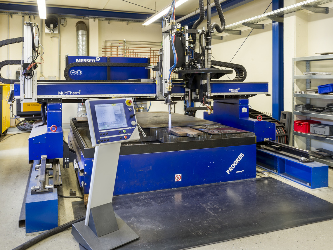 Große CNC-Schneidanlage der Marke Messer mit Bedienpult im Vordergrund steht in einer Werkhalle; das Gerät ist einsatzbereit, verschiedene Absaugvorrichtungen und Werkregale befinden sich im Hintergrund.