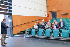 Elf ältere Personen, zehn Männer und eine Frau, sitzen in einem Hörsaal mit grünen Stühlen, vorne spricht ein Mann im Sakko stehend zur Gruppe