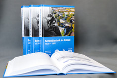 Mehrere aufgeschlagene Bücher liegen fächerförmig im Vordergrund, dahinter stehen Exemplare eines Buches mit dem Titel "Schweißtechnik im Grünen", blauem Cover und industriellem Motiv aufrecht vor neutralem Hintergrund 