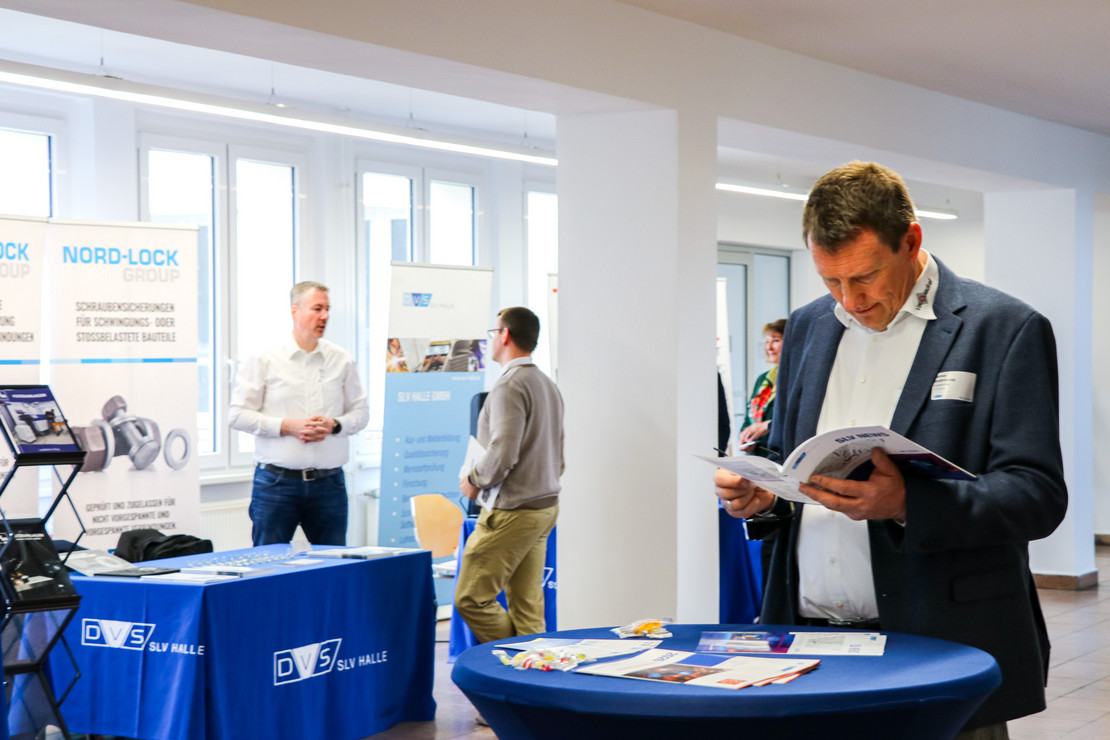 Person im Anzug liest die SLV-News, im Hintergrund ein Informationsstand mit Materialien und weitere Teilnehmende in einem Veranstaltungsraum
