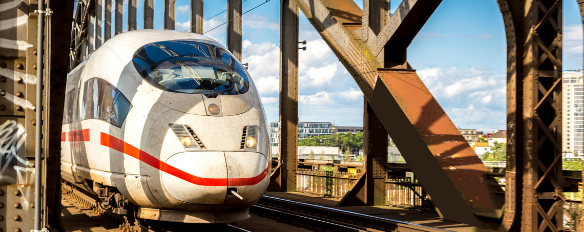 Ein weißer Fernzug mit rotem Streifen fährt über eine stählerne Eisenbahnbrücke bei Tageslicht; im Hintergrund sind Stadtgebäude und ein blauer Himmel mit Wolken zu sehen.