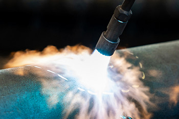 Nahaufnahme eines Flammrichtbrenners, der mit intensiver Flamme auf ein dickwandiges Stahlrohr trifft. Um die Schweißnaht herum entstehen sichtbare Hitzeverwirbelungen. Im Hintergrund sind unscharfe rote Schweißvorhänge und weitere große Metallbauteile erkennbar.