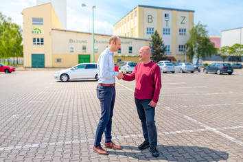 Zwei Männer stehen auf einem gepflasterten Platz und reichen sich die Hand. Im Hintergrund sind geparkte Autos und das BTZ Bernburg zu sehen.