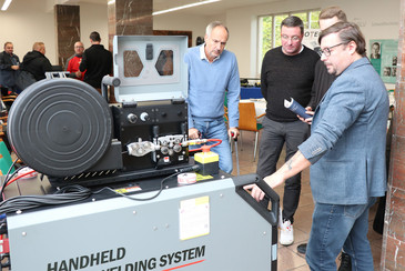 Ein Fachmann erklärt die Funktionen eines Handheld-Schweißsystems, umgeben von interessierten Teilnehmern.