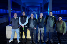 Fünf Männer und eine Frau stehen nebeneinander in einer großflächigen Halle mit dunklem Boden und blauer Beleuchtung, die architektonisch an ein leerstehendes Schwimmbecken oder ein unterirdisches Bauwerk erinnert.