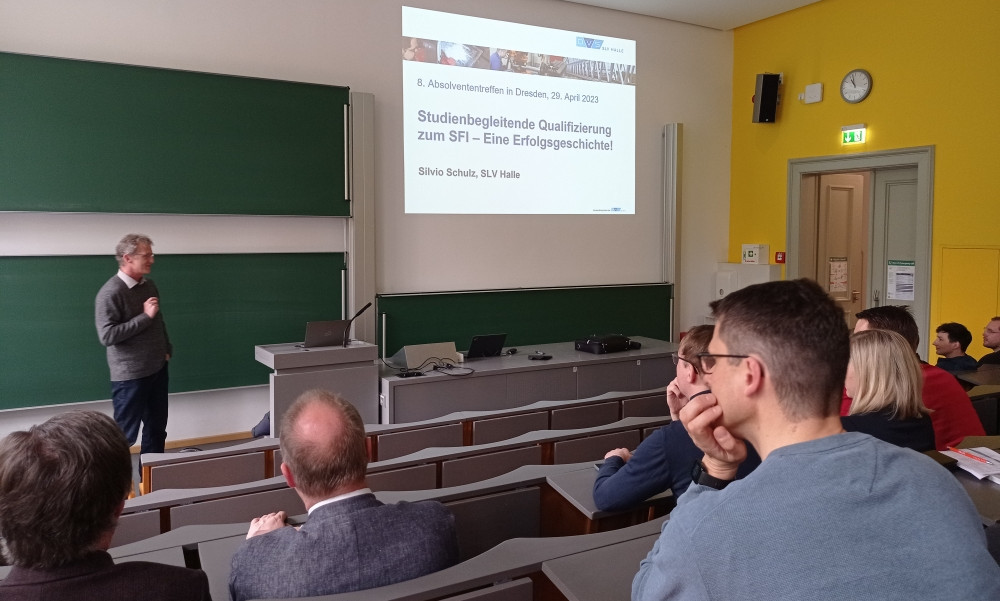 Ein Mann steht in einem Hörsaal vor einer Projektion mit dem Titel „Studienbegleitende Qualifizierung zum SFI – Eine Erfolgsgeschichte“ und spricht zu einem Publikum aus etwa zehn Personen, das aufmerksam zuhört.