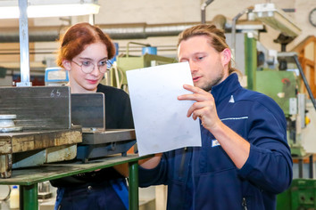 Ein Ausbilder und eine Auszubildende stehen in einer Werkstatt vor einer Maschine. Beide betrachten aufmerksam ein Werkstück und besprechen die Arbeitsschritte.