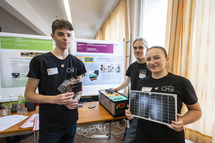 Drei junge Forschende in schwarzen GWG-T-Shirts präsentieren ihr Projekt. Einer hält Solarmodule, eine andere ein kleines Solarpanel, während der dritte an einer technischen Apparatur arbeitet. Im Hintergrund wissenschaftliche Poster mit Projektinformationen.