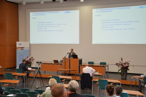 Ein Vortragender steht an einem Rednerpult in einem Konferenzsaal mit Leinwandprojektion, das Publikum sitzt mit Abstand in Reihen, während eine Kamera den Vortrag aufzeichnet.