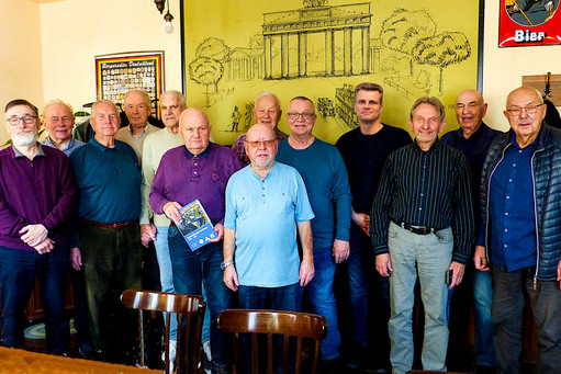 Gruppe älterer Männer steht dicht nebeneinander in einem Innenraum, einige halten kleine Geschenke oder Blumen, bunte Kleidung in Blau, Lila und Mustern, im Hintergrund Wanddekoration und ein Tisch 