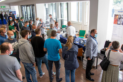 Zahlreiche Besucherinnen und Besucher mit Mundschutz stehen dicht gedrängt an mehreren Messeständen im Flur eines Konferenzgebäudes und informieren sich über technische Geräte.