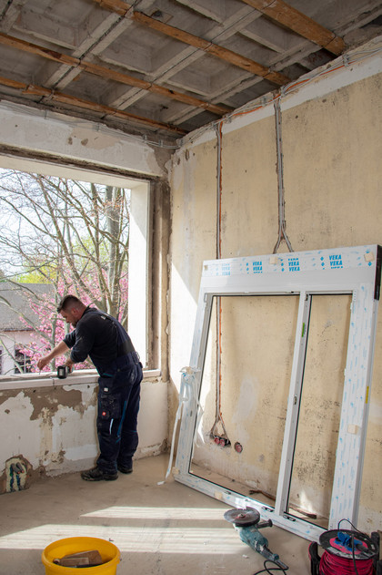 Ein Mann arbeitet in einem teilsanierten Raum an einem offenen Fenster, neben ihm lehnt ein neues Fensterrahmenelement an der Wand, Decke und Wände sind im Rohbauzustand, draußen blüht ein Baum.