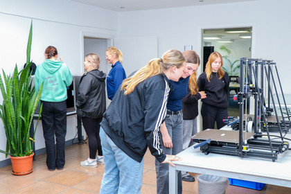 Mehrere Mädchen betrachten gemeinsam mit einer Betreuerin einen 3D-Drucker, im Hintergrund stehen weitere Jugendliche an einem Computerterminal.