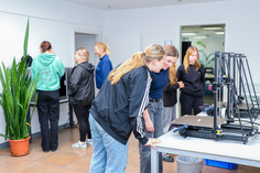 Mehrere Mädchen betrachten gemeinsam mit einer Betreuerin einen 3D-Drucker, im Hintergrund stehen weitere Jugendliche an einem Computerterminal.