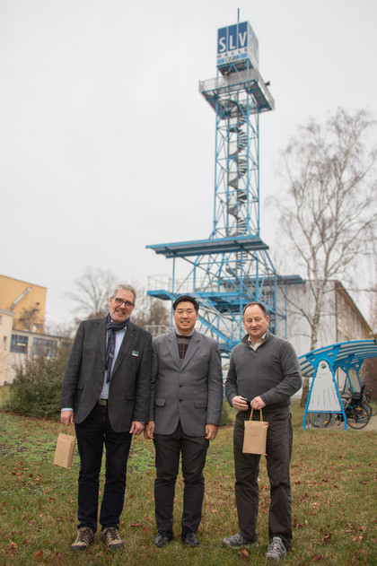 Drei Männer stehen mit Geschenktüten auf einer Wiese vor dem blauen SLV-Prüfturm in Halle, alle tragen Jacke oder Sakko, bewölkter Himmel im Hintergrund