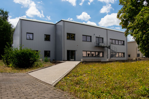 Modernes zweigeschossiges Industriegebäude mit grauer Metallfassade und Außentreppe, umgeben von Wiese, Bäumen und einem gepflasterten Weg, bei sonnigem Wetter aufgenommen.