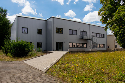 Modernes zweigeschossiges Industriegebäude mit grauer Metallfassade und Außentreppe, umgeben von Wiese, Bäumen und einem gepflasterten Weg, bei sonnigem Wetter aufgenommen.