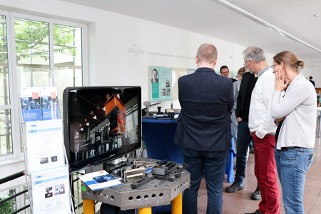 Vier Personen betrachten technische Exponate und Broschüren an einem Messestand, im Hintergrund Fenster und Infotafeln zur Geschichte der SLV Halle.