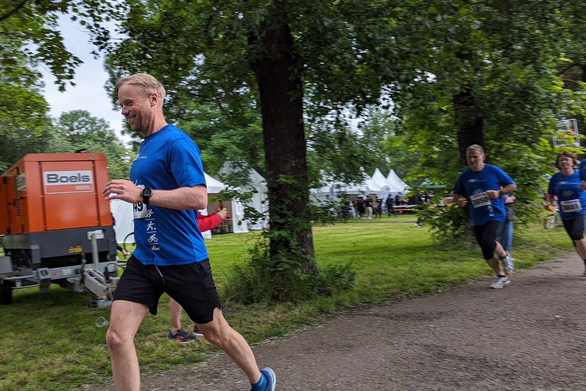 Ein Mann im blauen Laufshirt mit Startnummer läuft konzentriert auf einem Parkweg, gefolgt von weiteren Teilnehmenden des Firmenlaufs. Im Hintergrund stehen weiße Zelte, ein orangefarbenes Boels-Aggregat und mehrere Personen unter Bäumen.