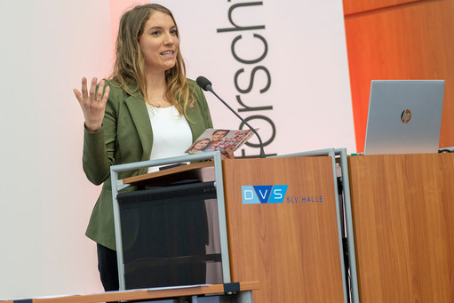 Eine Person steht an einem Rednerpult mit Mikrofon und spricht, während sie eine Broschüre in der Hand hält. Auf dem Pult steht ein Laptop, und das Logo „DVS SLV Halle“ ist sichtbar, im Hintergrund hängt ein Banner mit teilweise lesbarem Schriftzug.
