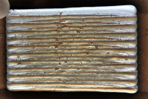 Nahaufnahme einer additiv gefertigten Metallprobe mit mehreren parallelen Schweißraupen; kleine Poren und Unregelmäßigkeiten sind zwischen den Schweißlagen sichtbar.