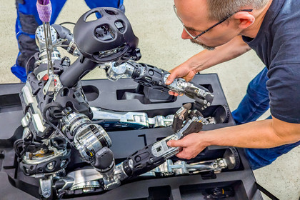 Ein Mann mit Brille hält die Arme eines sitzenden humanoiden Roboters, der ihn aus einer geöffneten Transportkoffer anblickt. Im Koffer liegen weitere metallische Roboterbauteile in Schaumstoffaussparungen.