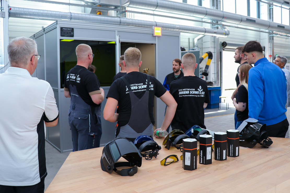 Jugendliche, auf deren T-Shirt Rückseite "Jugend schweißt" steht, erhalten eine technische Einführung durch eine Fachkraft in der Werkhalle, im Vordergrund Schutzausrüstung und Schweißhelme auf dem Tisch