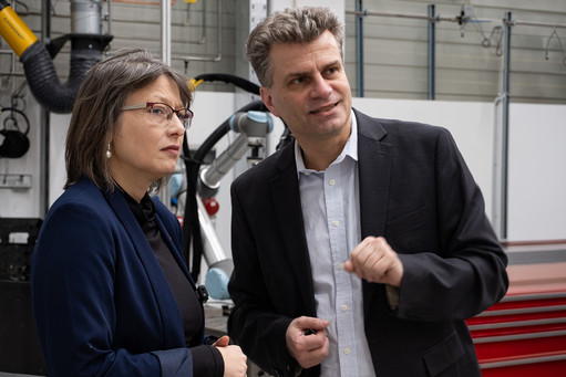 Die Landtagsabgeordnete Dr. Katja Pähle und Geschäftsführer Steffen Wagner stehen im Europäischen Berufsbildungszentrum Schweißen im Schienenfahrzeugbau. Beide tragen dunkle Kleidung, im Hintergrund sind Luftabzüge und Roboter zu sehen.