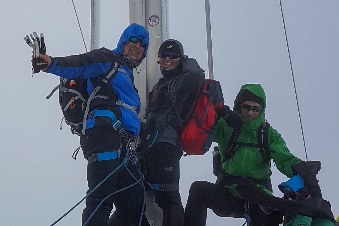 Drei Bergsteiger stehen auf einem Gipfel vor einem Gipfelkreuz. Sie tragen warme Outdoor-Kleidung mit Kapuzen und Rucksäcken. Das Wetter ist neblig, und sie lächeln in die Kamera.