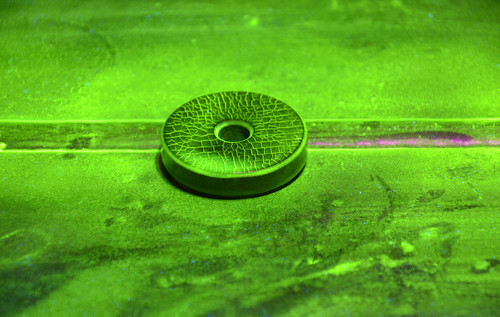 Eine runde Prüfscheibe mit feinen Rissstrukturen liegt auf einer metallischen Oberfläche, die mit fluoreszierendem Entwickler bedeckt ist. Die Darstellung erfolgt unter UV-Licht im Rahmen einer Farbeindringprüfung. Die feinen Risse sind deutlich sichtbar.