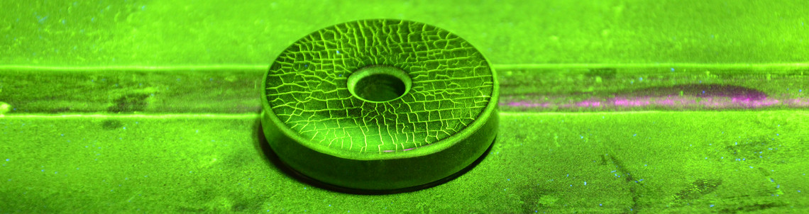 Eine runde Prüfscheibe mit feinen Rissstrukturen liegt auf einer metallischen Oberfläche, die mit fluoreszierendem Entwickler bedeckt ist. Die Darstellung erfolgt unter UV-Licht im Rahmen einer Farbeindringprüfung. Die feinen Risse sind deutlich sichtbar.