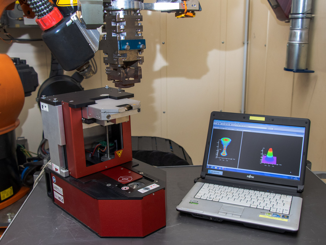 Ein Messgerät zur Analyse von Schweißnähten steht auf einem Tisch neben einem geöffneten Laptop, auf dessen Bildschirm zwei 3D-Auswertungen angezeigt werden; im Hintergrund ein Industrieroboterarm.