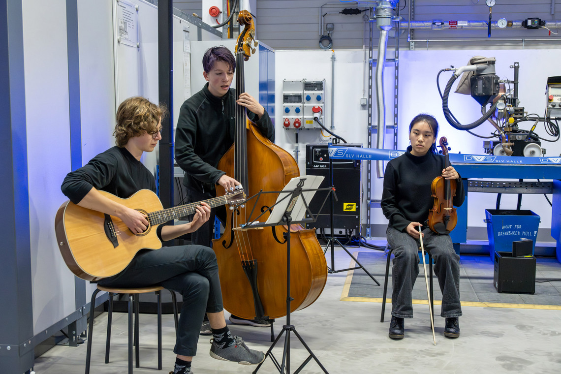 Drei junge Musikerinnen und Musiker in schwarzer Kleidung spielen in einer Werkhalle Gitarre, Kontrabass und Violine; im Hintergrund befinden sich Schweißtechnik, Maschinensteuerungen und blaue Beleuchtung.