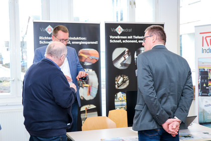 Drei Männer stehen an einem Messetisch vor Roll-Ups mit technischen Produktabbildungen, einer hält ein Gerät in der Hand und erklärt es den anderen beiden
