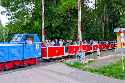 Offene Wagen einer rot-blauen Parkeisenbahn mit vielen Mitfahrenden passieren einen kleinen Haltepunkt im Grünen. Die Schranken sind oben.