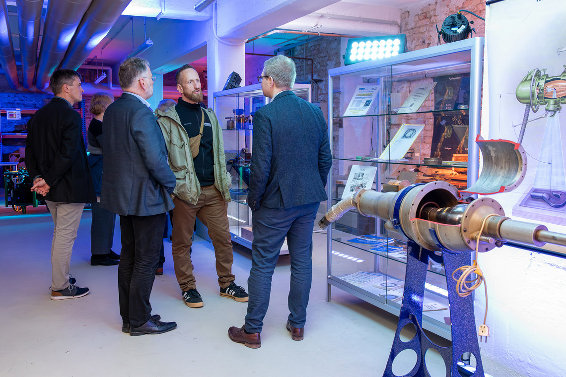 Mehrere Personen im Gespräch vor Vitrinen mit Exponaten und Fachliteratur in beleuchtetem Ausstellungsraum.