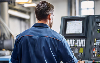 Mann in blauer Arbeitskleidung bedient ein Steuerpult mit Bildschirm in einer Industriehalle.