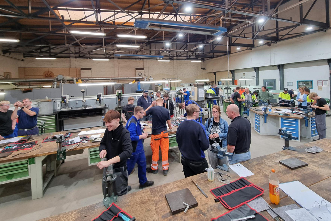 Mehrere Personen arbeiten in einer großen Werkhalle an verschiedenen Arbeitsplätzen mit Metallbauteilen und technischen Geräten. Im Vordergrund steht eine Gruppe um einen Arbeitsplatz, im Hintergrund sind weitere Personen, Tische und Arbeitsbereiche zu sehen.