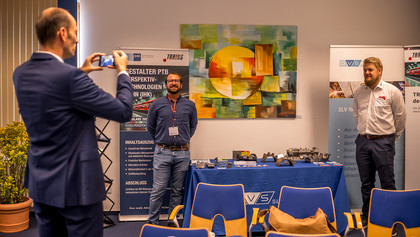 Zwei Männer stehen neben einem Messestand der SLV Halle in einem Innenraum. Auf einem blau gedeckten Tisch liegen Metallbauteile, Prüfstücke, Messgeräte und Informationsmaterial, daneben stehen Roll-up-Banner mit Bezug zu Bahntechnik und Schienenverkehr. Ein Mann fotografiert die beiden.