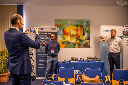 Zwei Männer stehen neben einem Messestand der SLV Halle in einem Innenraum. Auf einem blau gedeckten Tisch liegen Metallbauteile, Prüfstücke, Messgeräte und Informationsmaterial, daneben stehen Roll-up-Banner mit Bezug zu Bahntechnik und Schienenverkehr. Ein Mann fotografiert die beiden.