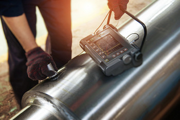 Prüfperson führt eine Ultraschallprüfung an einer Metallwelle durch, während die Messergebnisse auf einem tragbaren Gerät angezeigt werden.