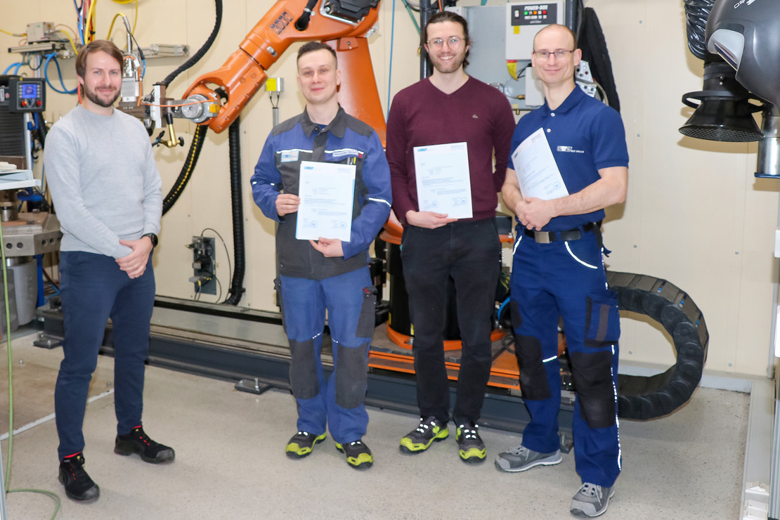 Drei Personen posieren mit Zertifikaten vor einer industriellen Roboterschweißanlage, links davon steht der Dozent. 