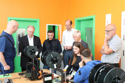 Sechs Männer stehen um einen Tisch mit mehreren 3D-Druckern, ein Mädchen und ein Junge bedienen ein Gerät, bunte Wände und grüne Türen im Hintergrund.