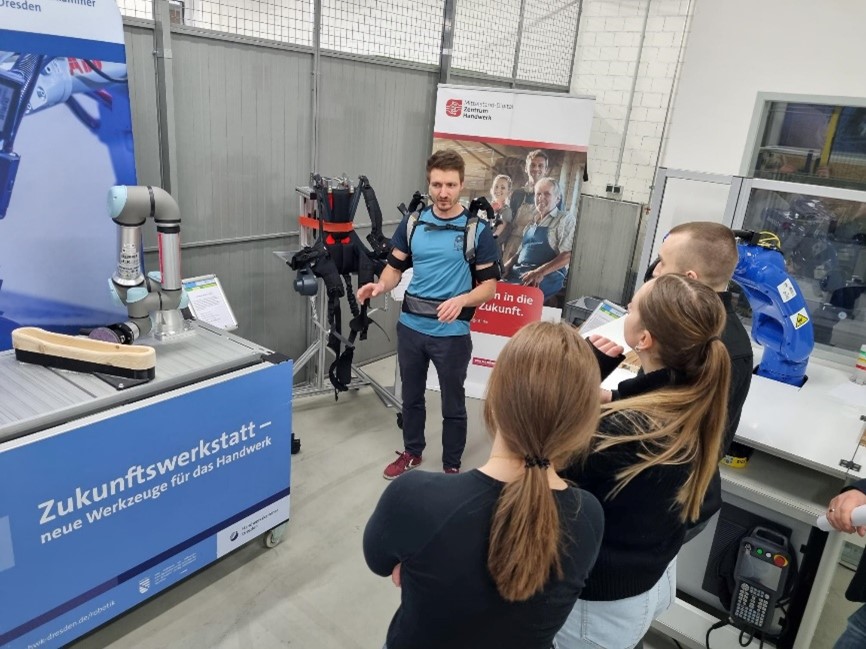 Ein Mann demonstriert vor einer kleinen Gruppe von Interessierten ein Exoskelett in einer Werkhalle; neben ihm steht ein Roboterarm und eine Arbeitsstation mit der Aufschrift „Zukunftswerkstatt – neue Werkzeuge für das Handwerk“.